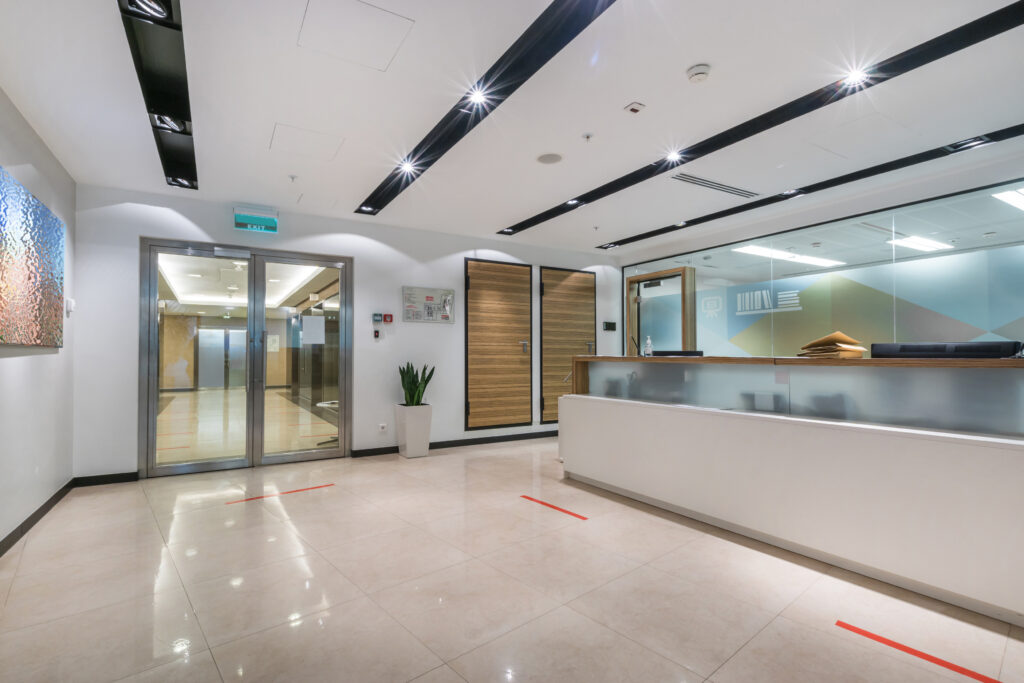 A commercial office hall with a reception desk, glass doors and a picture on the wall. Red line markings for social distance